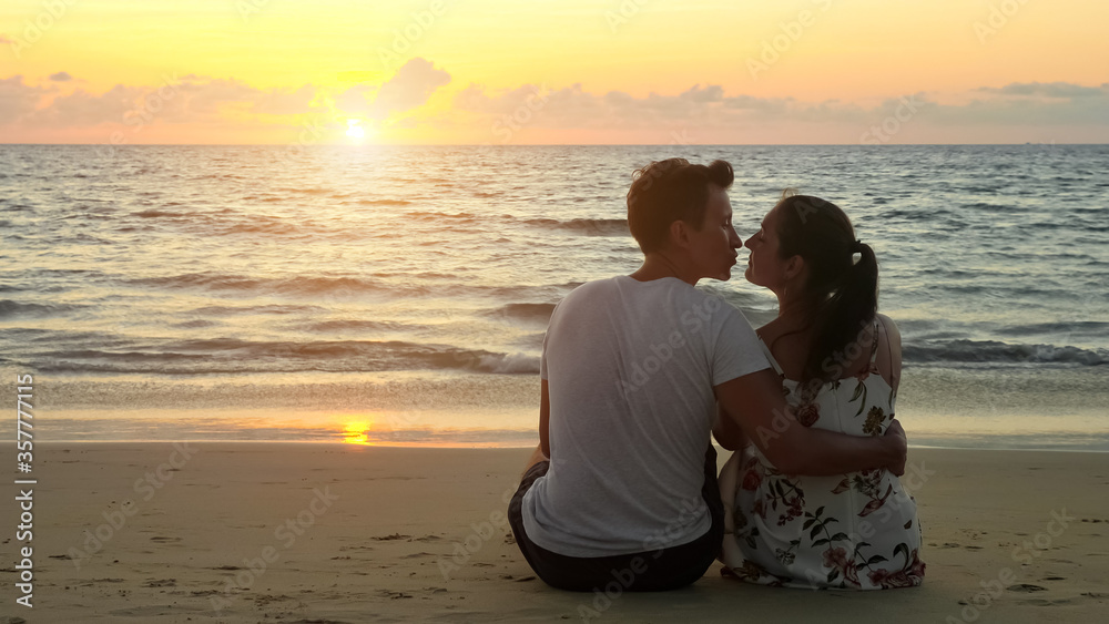 lovely young couple hugs and kisses sitting on empty ocean beach watching sunset at tropical resort in evening backside view