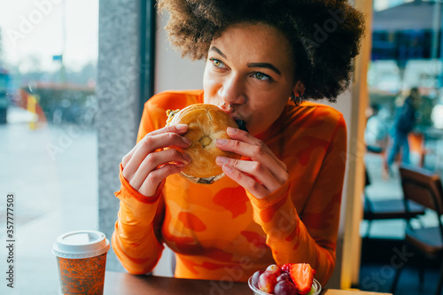 Young beautiful afro woman indoors restaurant biting hamburger - hungry diverse woman sitting restaurant eating bagel