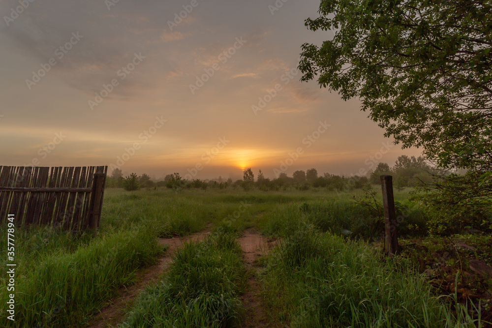 Sun is rising over the beautiful and picturesque field with old wooden fence and gravel road. Horizontal landscape photography.