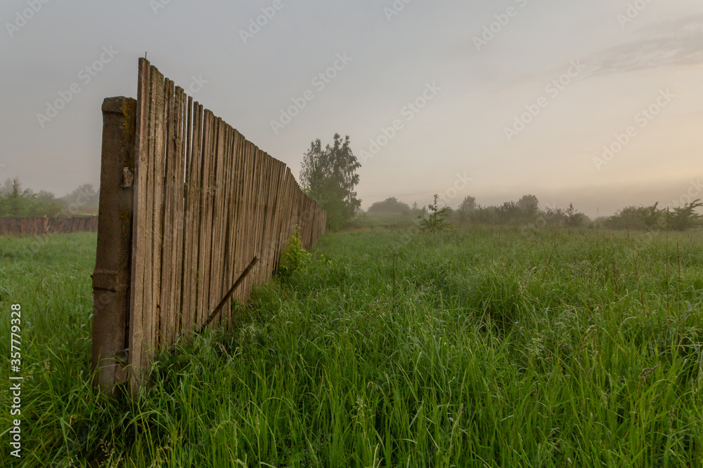 Scenic panorama with wooden fence in the grass field in early misty morning.