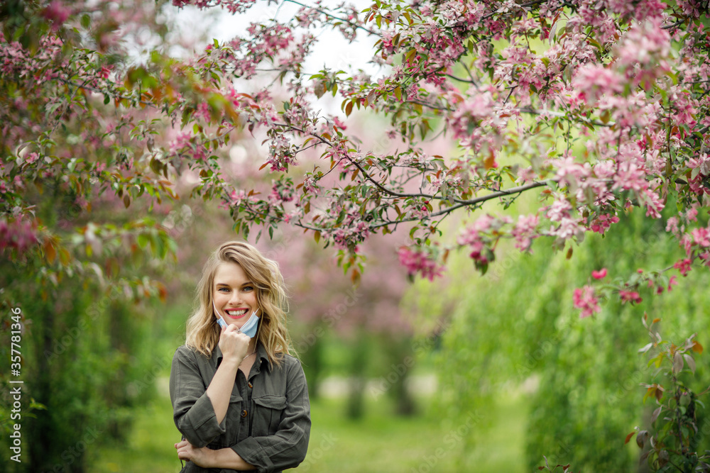 Young woman in medical mask on background of flowering trees in park at afternoon. Young woman in medical mask on background of flowering trees in park at afternoon.