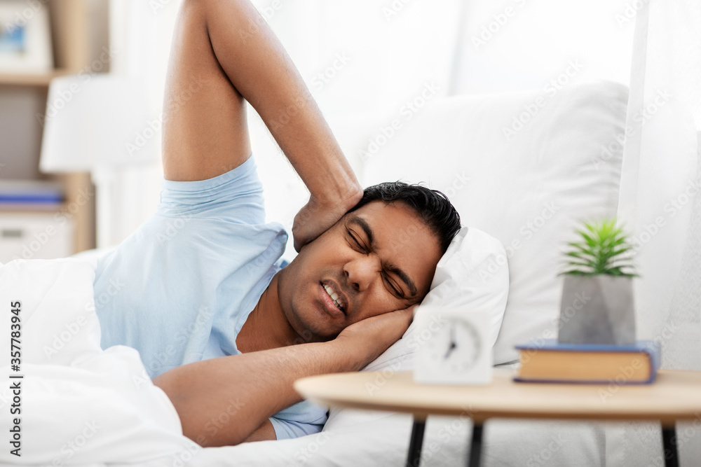people, bedtime and rest concept - annoyed indian man with alarm clock lying in bed at home and covering ears people, bedtime and rest concept - annoyed indian man with alarm clock lying in bed at home and covering ears