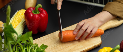 Female cook slicing fresh carrot on chopping block at dark modern kitchen