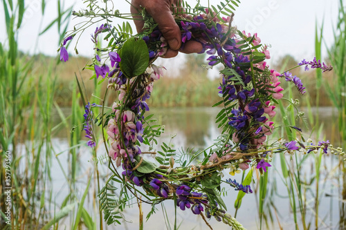  Multicolored wreath of wildflowers in human hand on lake background in Ukraine.