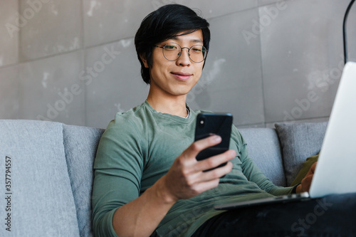 Image of pleased asian man using cellphone and laptop while sitting