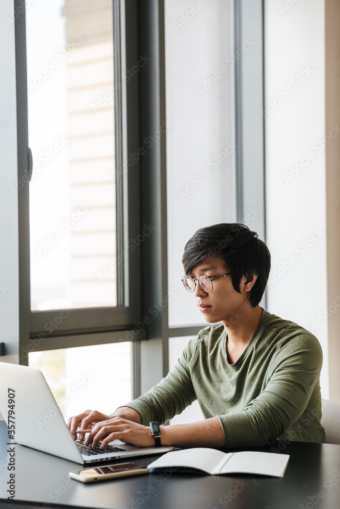 Image of serious asian man working with laptop while sitting at table Image of serious asian man working with laptop while sitting at table