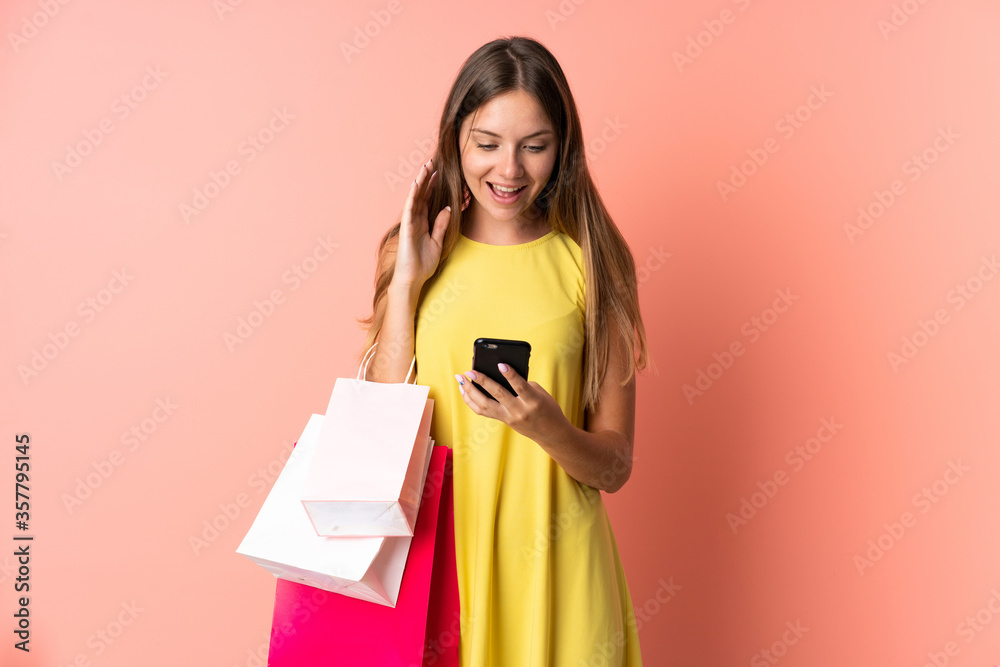 Young Lithuanian woman isolated on pink background holding shopping bags and writing a message with her cell phone to a friend