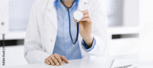 Unknown woman-doctor in blue blouse is holding stethoscope head while sitting at the desk in clinic  close-up. Physician ready to examine and help patient. Medicine concept