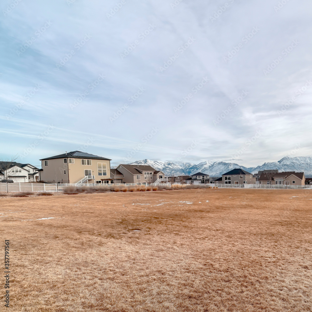 Square crop Grassy field with melting snow against homes and snowy mountain background Square crop Grassy field with melting snow against homes and snowy mountain background