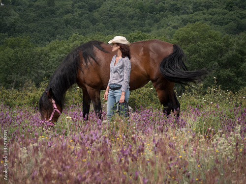 Horizontal view of female horse coach with white hat looking at one side and andalusian horse grazing in a spring meadow.