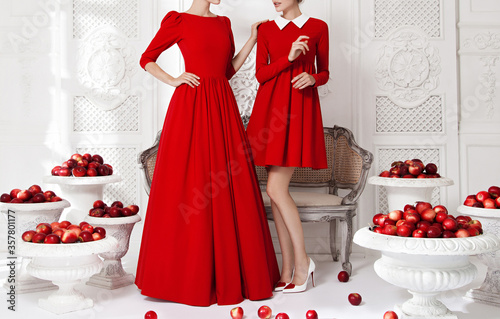 Two beautiful young girls models, posing in luxurious red dresses in the studio, amid scenery with openwork moldings, a carved sofa, white vases with red apples