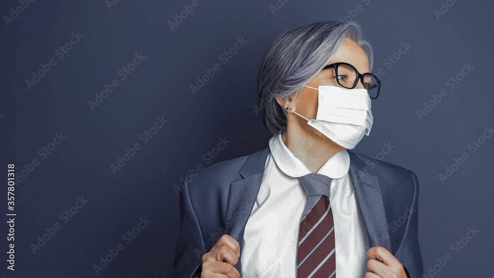 Mature woman wearing mask corrects her jacket looking at right side. Caucasian smart business woman on gray background. Copy space at left. Quarantine concept. Toned image.