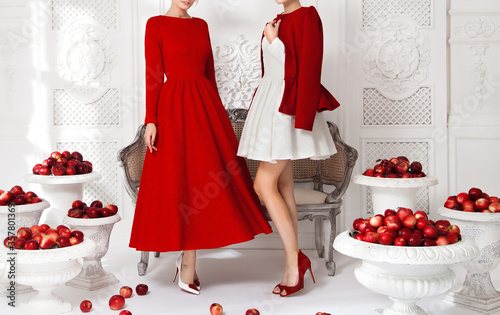 Two beautiful young girls models, posing in luxurious white and red dresses in the studio, amid scenery with openwork moldings, a carved sofa, white vases with red apples