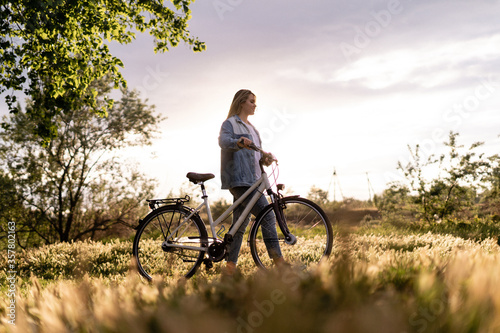 A young woman is walking near a bicycle in a city park at summer sunset. Blurred background