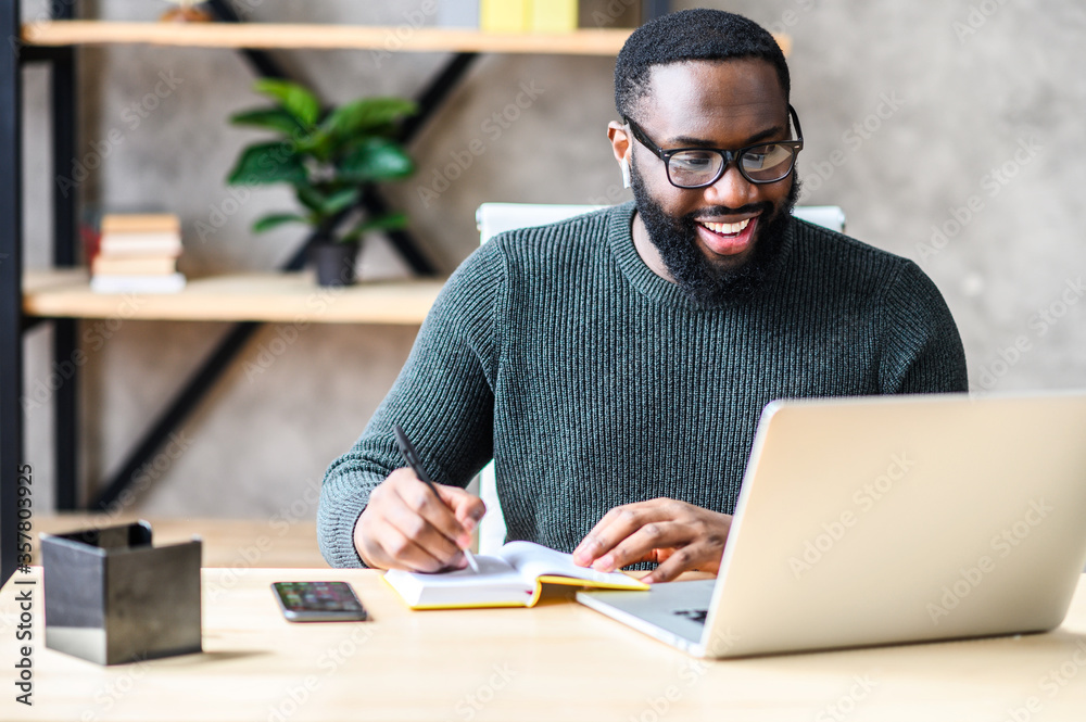 Cheerful African-American male student or worker in glasses is watching online lectures or webinars and writing notes in a notebook Cheerful African-American male student or worker in glasses is watching online lectures or webinars and writing notes in a notebook