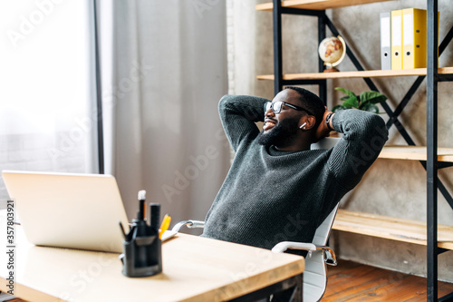 The young black guy sits at the desk in the office, he leaned back in his chair relaxed. End of good business day