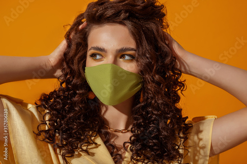 Fashionable curly woman wearing trendy outfit with green protective face mask. Summer fashion during quarantine of coronavirus outbreak. Model posing on orange background