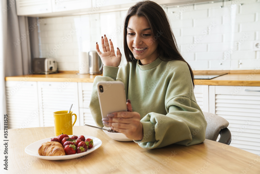 Photo of woman using mobile phone and waving hand while having breakfast