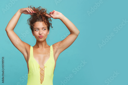 Studio photo of positive young lovely red haired curly woman keeping hands above her head and pouting lips while standing over blue background in yellow swimsuit