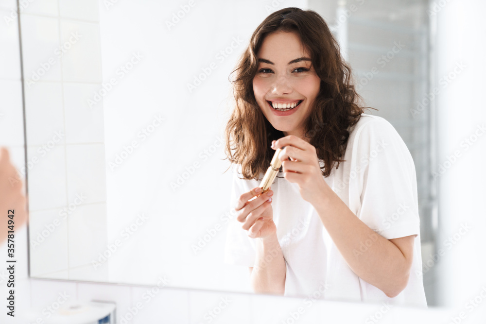 Photo of woman smiling and holding mascara while looking at mirror Photo of woman smiling and holding mascara while looking at mirror