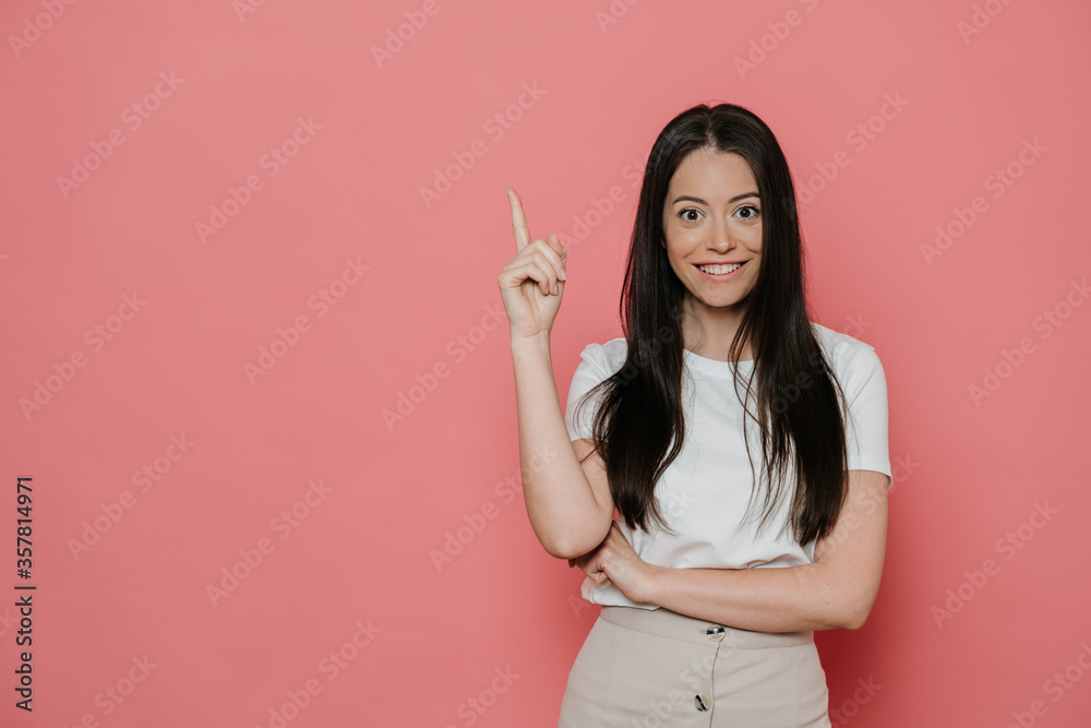 Cheerful pretty brunette in white t-shirt, beige skirt, pointing upward by index finger, in amazed mood, enjoying her life, ready for summertime. Stands over pink backdrop. Sale and promotion concept. - obrazy, fototapety, plakaty 