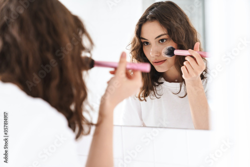 Photo of serious woman using powder brush while looking at mirror