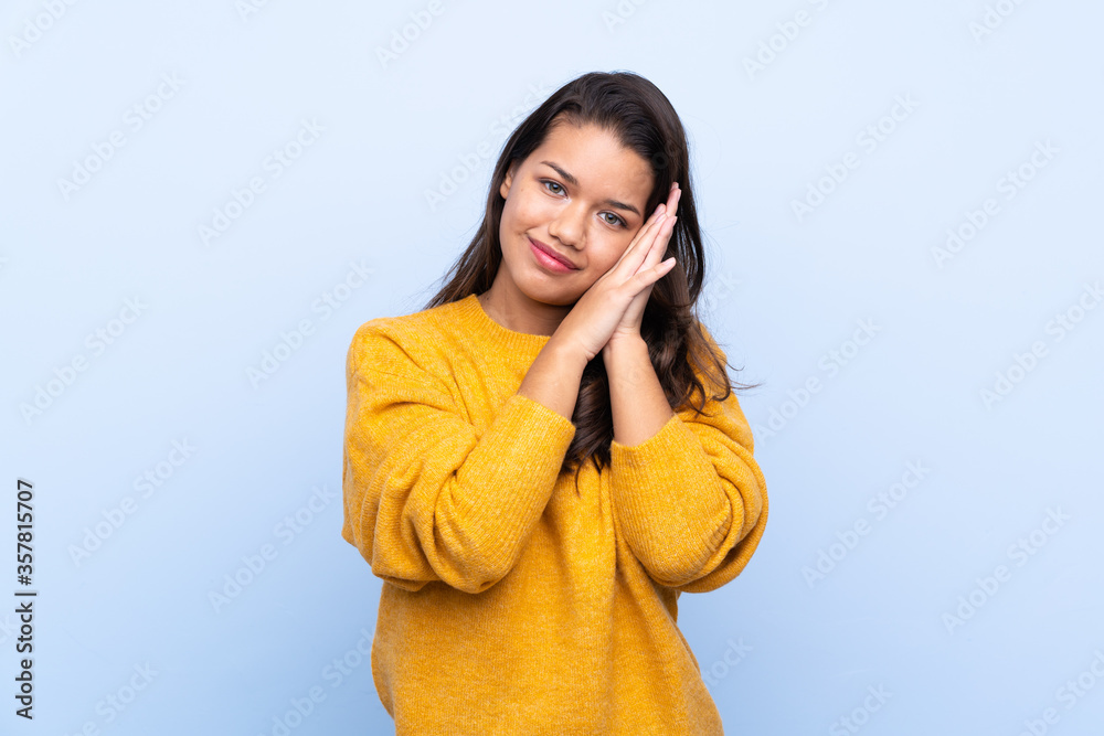 Young Colombian girl with sweater over isolated blue background making sleep gesture in dorable expression Young Colombian girl with sweater over isolated blue background making sleep gesture in dorable expression