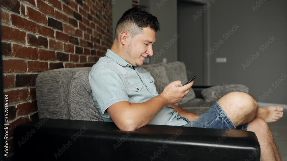 A man with a black smartphone in his hands sits on a sofa in a living room. He smiles and laugh. He browsing and texting. A man is wearing in summer jeans clothes A man with a black smartphone in his hands sits on a sofa in a living room. He smiles and laugh. He browsing and texting. A man is wearing in summer jeans clothes