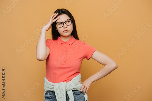 Thoughtful displeased woman with, holds chin and looks away, contemplates about something, wears pink t shirt, being in upset mood, smirks face and being upset by bad thoughts over yellow backdrop.