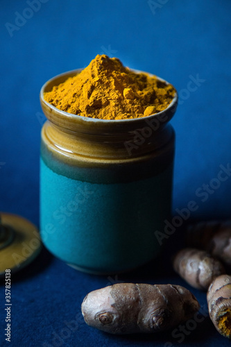 Turmeric Powder in a Ceramic Jar surrounded by Haldi Roots on a Blue Background. Yellow Southeast Asian Spice also known as Curcuma or Saffron Des Indes.