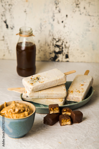 Snickers Popsicles stacked on top of other surrounded by Chocolate Pieces and Caramel Syrup. Chocolate Icecream Bars, white background with soft focus.