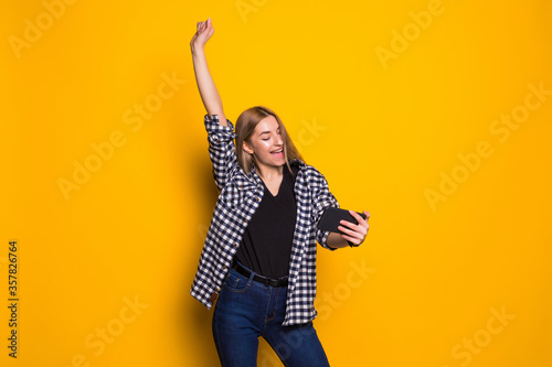 Portrait of a cheerful young woman holding mobile phone  celebrating standing isolated over yellow background