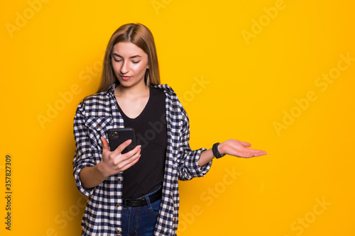 Young woman wearing basic clothes expressing discontent while holding cellphone isolated over yellow background