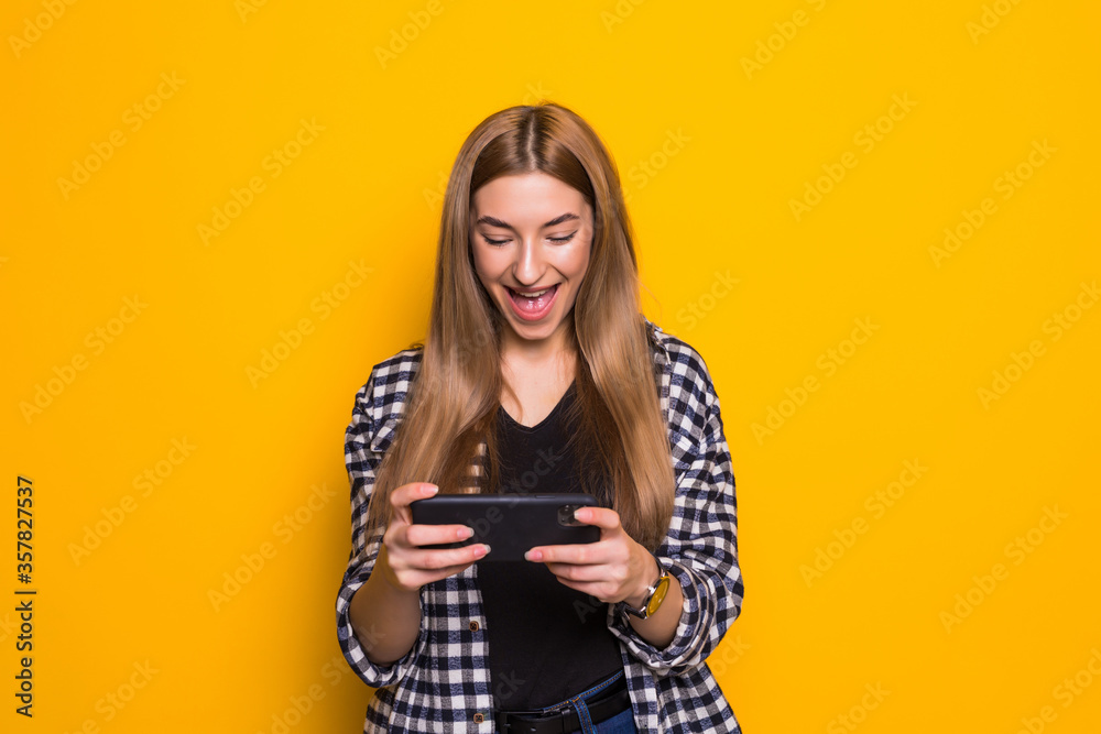Young happy cute beautiful young woman play games by mobile phone isolated over yellow background. Young happy cute beautiful young woman play games by mobile phone isolated over yellow background.