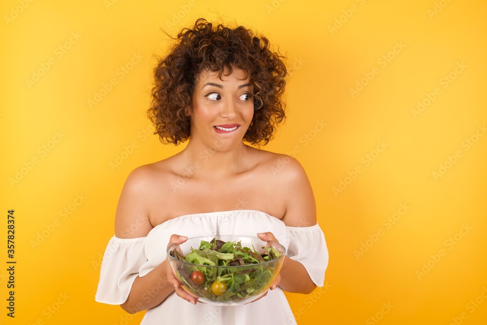 Dreamy young female with thoughtful expression, looks away keeps hands down bitting her lip thinks about something pleasant poses against gray wall. Dreamy young female with thoughtful expression, looks away keeps hands down bitting her lip thinks about something pleasant poses against gray wall.