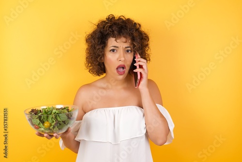 Young businesswoman holding a salad talking on the phone over isolated background stressed with hand on face, shocked with shame and surprise face, angry and frustrated. Fear and upset for mistake.