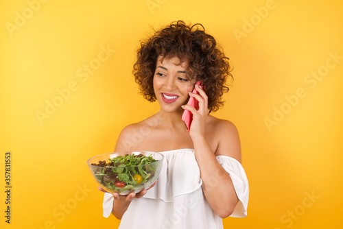 Lifestyle Concept - Portrait of beautiful Caucasian joyful woman holding a salad talking on mobile phone with friend. Yellow pastel studio background. Copy Space.