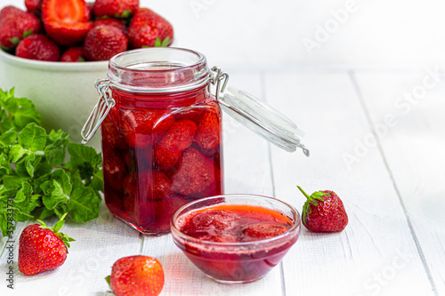 Strawberry jam in a glass jar next to fresh strawberries. On a white wooden background. Homemade winter fruit blanks. Selective focus.