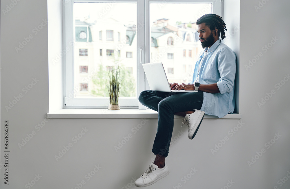 Thoughtful young African man using laptop while sitting on the window sill indoors Thoughtful young African man using laptop while sitting on the window sill indoors