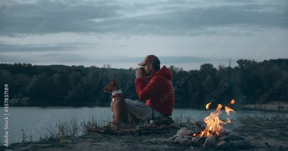 Man and pet dog rest next to campfire on hike trip. High quality 4k footage