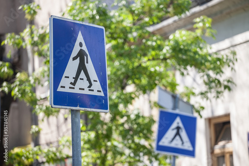 Two roadsigns warning drivers of the presence of a crosswalk with pedestrians crossing in an urban landscape, abiding by the European standards, with its typical blue template.