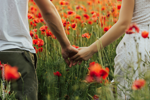 Obraz na płótnie Close up outdoor portrait of couple holding hands in the red poppy field. Love - romantic couple holding hands. . Young woman and man in love walking hand in hand on field. w salonie