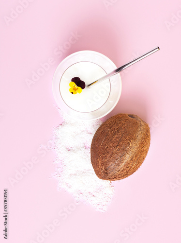 Coconut milk in a glass cup on a pink background, coconut lies next to it. Coconut milk is decorated with an edible violet flower. Top view, vertical orientation.