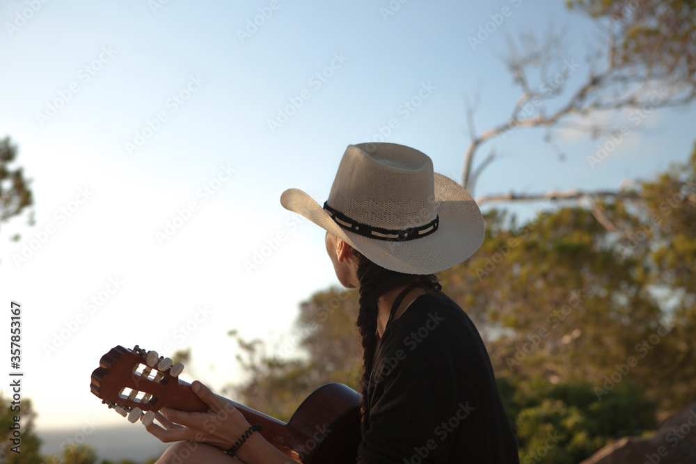 Wanderlust concept o woman with a cowboy look traveling with her guitar, the girl is wearing a hat and the evening light shines in the sky. The woman has views of the Mediterranean Sea Majorca Island