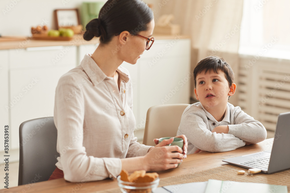 Warm-toned portrait of young mother talking to son while doing homework together sitting at table in cozy kitchen interior, copy space Warm-toned portrait of young mother talking to son while doing homework together sitting at table in cozy kitchen interior, copy space