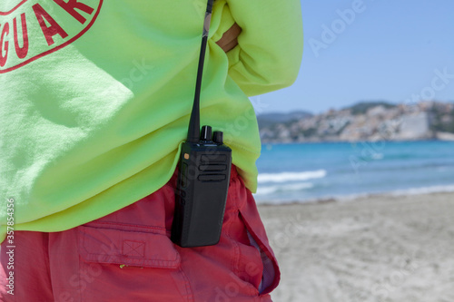 A male lifeguard on the beach during the summer. The man is wearing a sweatshirt and has a walkie talkie hanging on his swimsuit. Summer in Mallorca