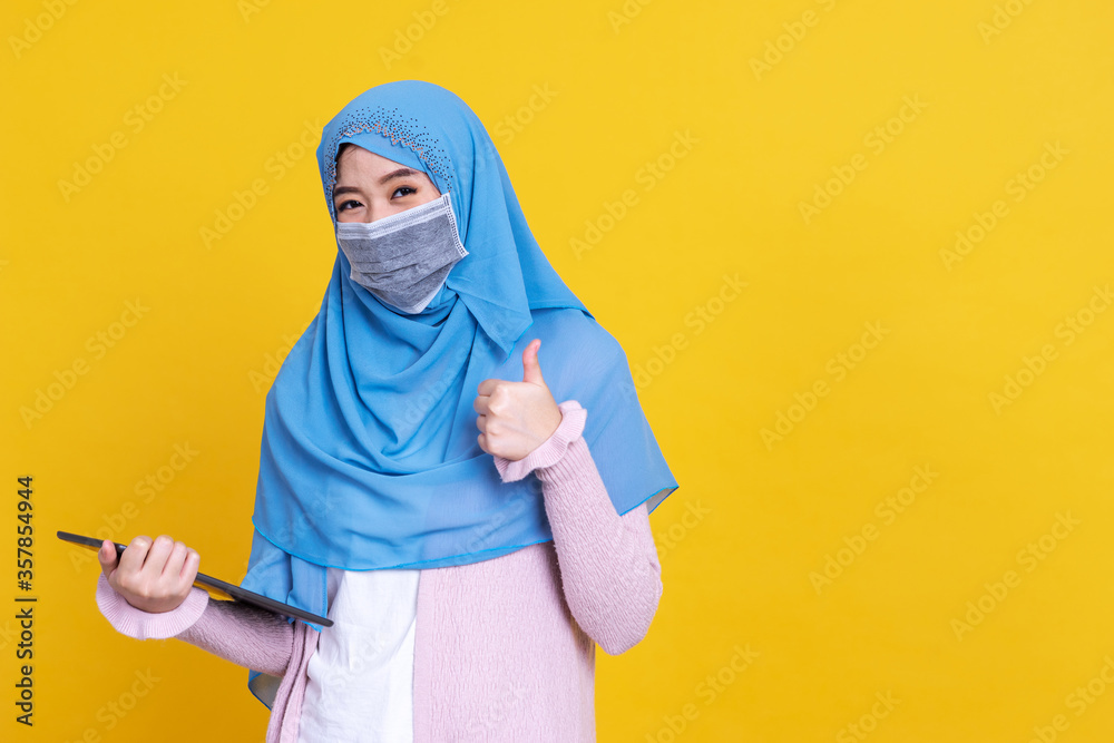 Asian Muslim woman wearing medical mask in hijab and using digital tablet isolated on color background Asian Muslim woman wearing medical mask in hijab and using digital tablet isolated on color background