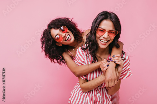 Young, spectacular models vigorously pose for portrait in studio. Tanned sisters embrace and laugh in unusual glasses photo