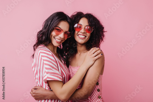 Excited african female model in round sunglasses embracing best friend. Indoor photo of cheerful black young woman enjoying photoshoot in studio