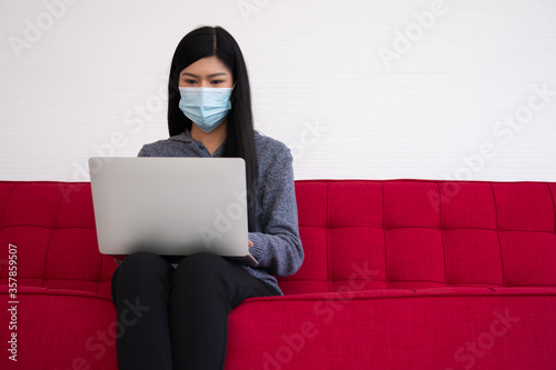 Asian woman wearing a face mask and using a laptop on the sofa for working from home. New lifestyle normal during a quarantine. Concept of stay home.